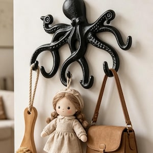 May include: A black octopus-shaped wall hook with multiple curved tentacles serving as individual hooks. The octopus is mounted on a wall and holds a wooden brush with a rope loop, a small fabric doll in a beige dress, and a brown leather-like handbag.