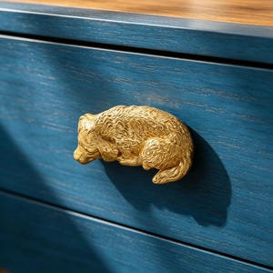 May include: A gold-colored bear-shaped drawer pull mounted on a blue wooden drawer. The bear is in a resting position, with detailed fur texture. The drawer is part of a larger piece of furniture with a wooden top.