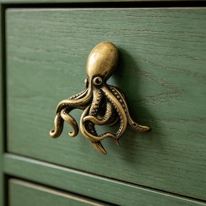 Könnte beinhalten: Ein messingfarbener Schubladengriff in Form eines Oktopus mit detaillierten Tentakeln und strukturierter Oberfläche, montiert an einer grünen Holzschubladenfront mit sichtbarer Maserung. Die Oktopusfigur hat einen glatten Kopf und Saugnäpfe.
