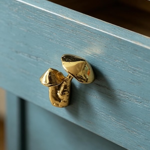 May include: A polished gold-toned metal cabinet knob shaped like two mushrooms of varying sizes, attached to a blue-painted wooden drawer front with a visible wood grain texture and an open top edge showing the interior space.