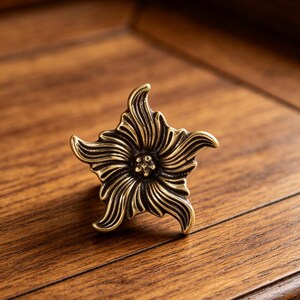 Puede incluir: Un tirador de caj&oacute;n de metal en tono lat&oacute;n con forma de flor de cinco p&eacute;talos. La superficie presenta surcos profundos y curvos que irradian desde un punto central, creando una apariencia texturizada y dimensional con reflejos oxidados.