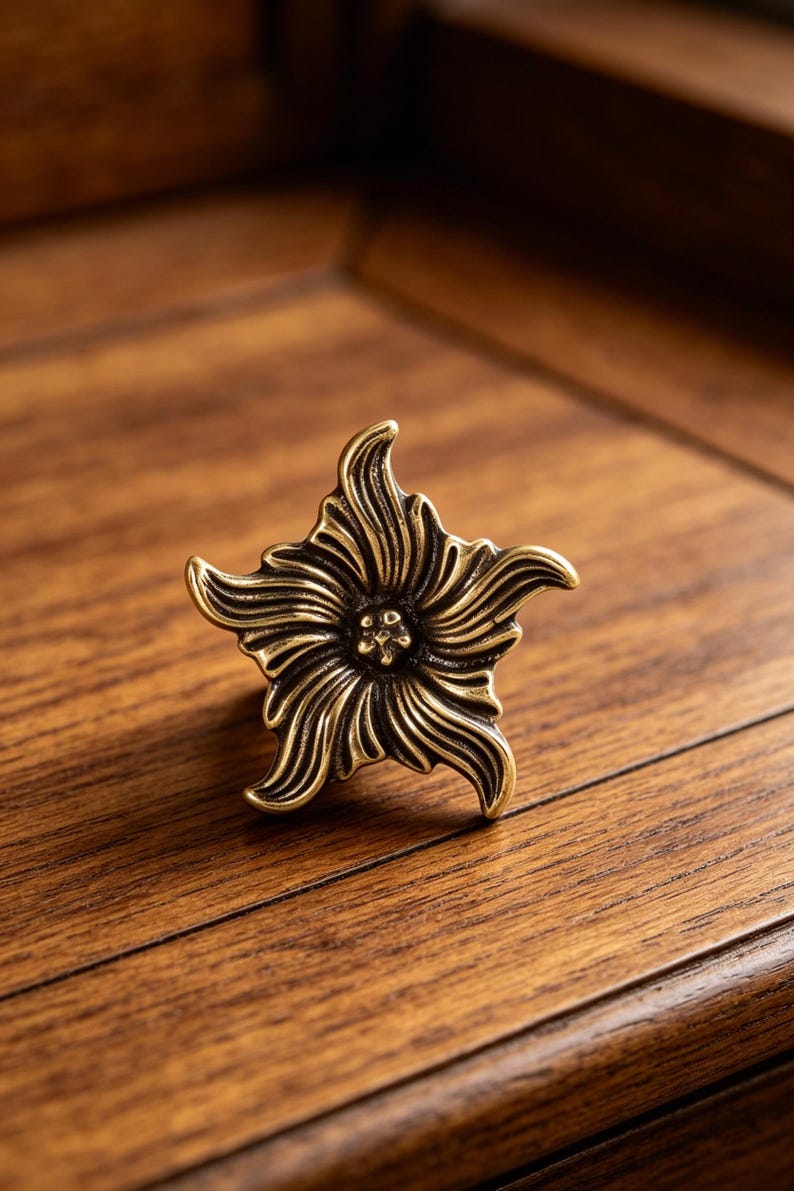 Puede incluir: Un tirador de caj&oacute;n de metal en tono lat&oacute;n con forma de flor de cinco p&eacute;talos. La superficie presenta surcos profundos y curvos que irradian desde un punto central, creando una apariencia texturizada y dimensional con reflejos oxidados.
