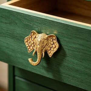 May include: A dark green wooden drawer featuring a metallic gold-coloured elephant-shaped handle. The elephant handle displays intricate textured details on its ears and trunk, mounted centrally on the front panel of the open drawer unit.