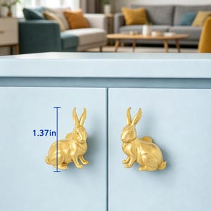 Puede incluir: Dos pomos de armario con forma de conejo de color dorado montados en una puerta de armario azul claro. Cada pomo presenta una figura de conejo detallada en posici&oacute;n sentada. Una l&iacute;nea gr&aacute;fica azul indica una altura de 3,48 cm.