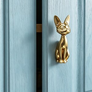 Op de afbeelding: Een gepolijste goudkleurige metalen kastknop in de vorm van een zittende kat met puntige oren en grote ogen. De knop is bevestigd op een lichtblauwe houten kastdeur met een zichtbare houtnerfstructuur en een verticaal paneelontwerp.