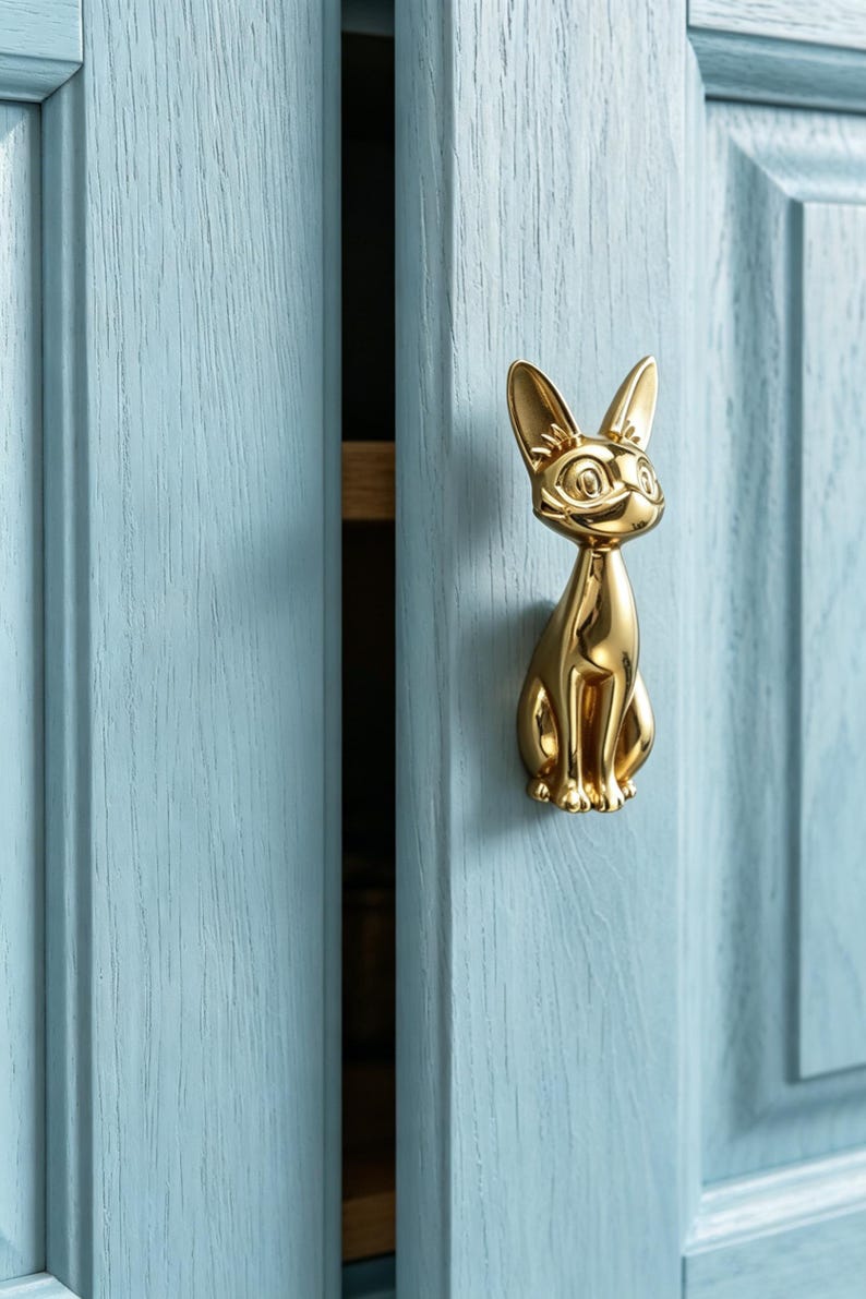 Op de afbeelding: Een gepolijste goudkleurige metalen kastknop in de vorm van een zittende kat met puntige oren en grote ogen. De knop is bevestigd op een lichtblauwe houten kastdeur met een zichtbare houtnerfstructuur en een verticaal paneelontwerp.