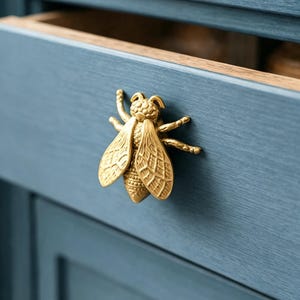 Puede incluir: Un tirador de armario dorado en forma de abeja está montado en una puerta de armario azul. El tirador presenta alas y cuerpo texturizados detallados. La puerta del armario está pintada con un acabado azul mate, con veta de madera visible.