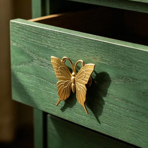 Könnte beinhalten: Eine dunkelgrüne Holzschublade mit einem markanten goldfarbenen Metallgriff in Schmetterlingsform. Die Schubladenfront zeigt eine sichtbare Holzmaserung, und der Schmetterlingsbeschlag ist mittig platziert und weist detaillierte Flügelmuster auf.