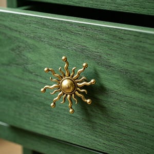 Op de afbeelding: Een donkergroen houten ladefront met een opvallende messingkleurige metalen knop in de vorm van een zon, met een centrale ronde koepel en uitstralende golvende stralen die eindigen in kleine bolletjes, met een zichtbare houtnerf op het oppervlak.