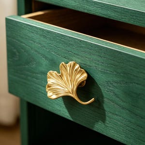 Puede incluir: Primer plano de una mesita de noche de madera verde con un cajón abierto. El tirador del cajón es una hoja de ginkgo dorada. La mesita de noche tiene una textura de veta de madera visible.