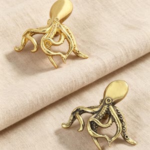 May include: Two gold-toned metal cabinet knobs shaped like octopuses. Each knob features a smooth, rounded head and detailed, curved tentacles with visible suction cup textures. The metallic finish has a slight sheen, highlighting the intricate, realistic form of the sea creature design on each individual piece.