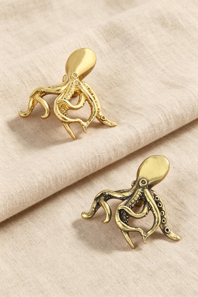 May include: Two gold-toned metal cabinet knobs shaped like octopuses. Each knob features a smooth, rounded head and detailed, curved tentacles with visible suction cup textures. The metallic finish has a slight sheen, highlighting the intricate, realistic form of the sea creature design on each individual piece.