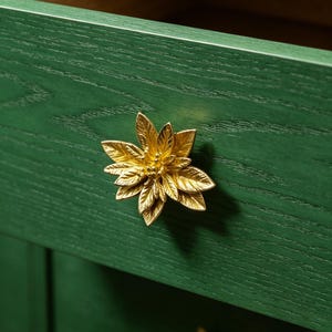 May include: A dark green wooden drawer featuring a metallic gold cabinet knob shaped like a multi-layered flower with detailed leaves. The drawer is slightly open, revealing a light wood interior, and a second identical gold knob is visible below.