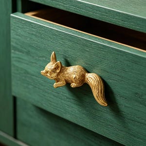 May include: A gold-colored squirrel-shaped drawer pull is mounted on a dark green wooden drawer. The drawer is partially open, revealing a light brown interior. The pull is detailed and adds a whimsical touch to the furniture.