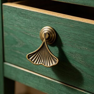 Puede incluir: Primer plano de un cajón de madera verde con un tirador en forma de hoja de ginkgo de color latón. El cajón está abierto, revelando un interior claro. La veta de la madera es visible, añadiendo textura.