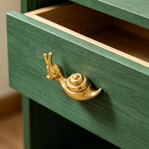 May include: A green wooden nightstand with a drawer slightly open, revealing a light brown interior. The drawer pull is a gold-colored snail. The nightstand has a simple, modern design.
