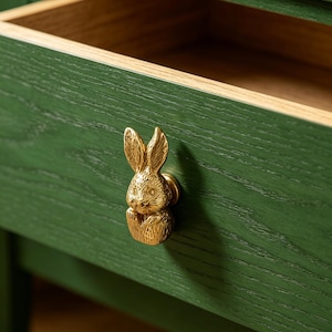 May include: A dark green wooden drawer featuring a gold-toned metal rabbit-shaped knob. The drawer is partially open, revealing the natural light-colored wood interior. The exterior surface of the drawer displays a visible wood grain texture.