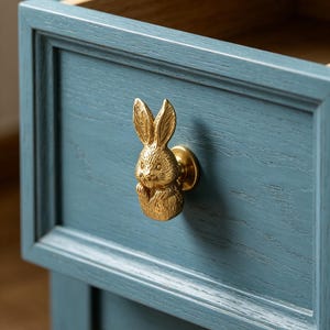 May include: A gold-toned metal cabinet knob shaped like a rabbit with textured fur details. The knob is attached to a blue-painted wooden drawer front with a visible wood grain texture and a recessed panel design on the drawer face.