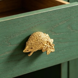 Op de afbeelding: Een goudkleurige egelvormige trekker is gemonteerd op een groene houten lade. De lade is gedeeltelijk open en onthult een lichtbruin interieur. De egel trekker heeft gedetailleerde kenmerken en is met schroeven aan de lade bevestigd.