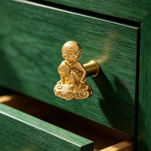 Puede incluir: Un pomo de mueble de metal dorado con forma de mono sentado sobre una nube, fijado a un cajón de madera verde oscuro. La figura del mono presenta detalles de pelaje texturizado y rasgos faciales, montada sobre una base cilíndrica.