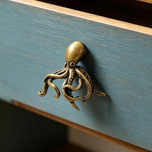 May include: A brass-toned metal drawer pull shaped like an octopus with detailed tentacles and a smooth head, mounted on a blue-painted wooden drawer front with a visible wood grain texture and a natural wood interior edge.