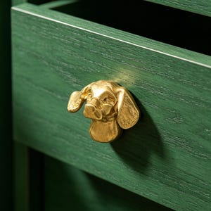 May include: A gold-toned metal cabinet knob shaped like the head of a dog with long, floppy ears. The knob is mounted on the front of a dark green wooden drawer, featuring a visible wood grain texture and a horizontal design.