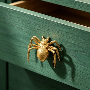 May include: A gold-colored spider-shaped drawer pull is mounted on a dark green wooden drawer. The drawer is partially open, revealing a light brown interior. The spider pull adds a unique, decorative touch to the furniture.