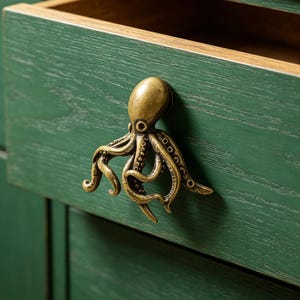 May include: A brass-toned metal octopus-shaped drawer pull attached to a green wooden drawer front. The octopus features detailed tentacles and a smooth head, providing a functional handle for opening the drawer on the green cabinet unit.