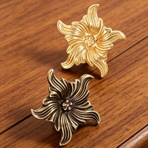 Puede incluir: Dos pomos de metal para cajones con forma de flores de cinco p&eacute;talos. Un pomo tiene un acabado dorado brillante y pulido, mientras que el otro presenta un acabado de bronce antiguo m&aacute;s oscuro. Ambos muestran texturas detalladas en relieve.