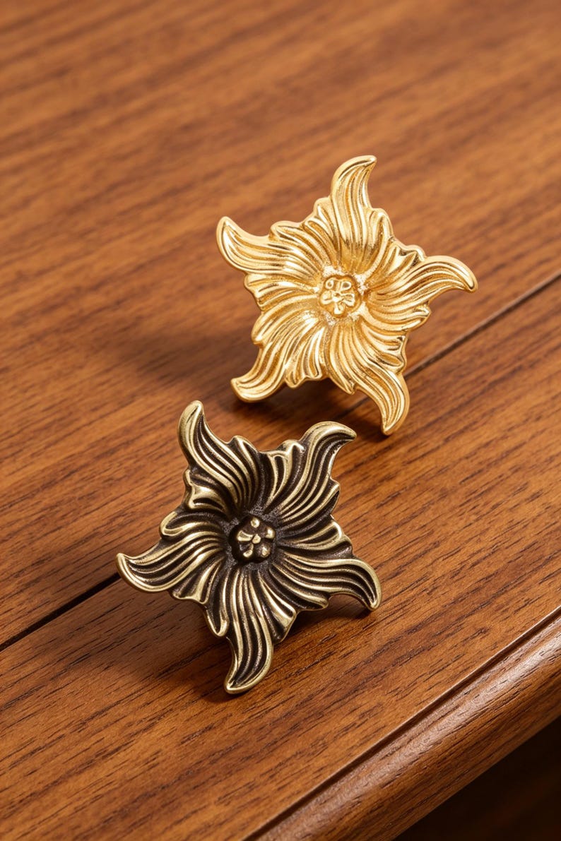 Puede incluir: Dos pomos de metal para cajones con forma de flores de cinco p&eacute;talos. Un pomo tiene un acabado dorado brillante y pulido, mientras que el otro presenta un acabado de bronce antiguo m&aacute;s oscuro. Ambos muestran texturas detalladas en relieve.