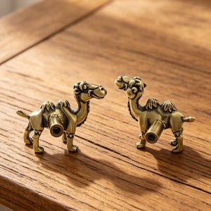 May include: Two brass-toned metal cabinet knobs shaped like camels. Each knob features a detailed camel figure with a protruding cylindrical mounting post on the side, finished in an antique gold color with dark recessed accents for depth.
