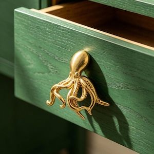 May include: A gold-toned metal octopus-shaped drawer pull attached to a dark green wooden drawer. The octopus features detailed tentacles and a smooth head, serving as a functional handle for the partially open drawer with visible wood grain.