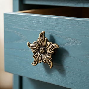 Puede incluir: Un caj&oacute;n de madera azul cerceta con un pomo de metal prominente en forma de estrella de cinco puntas con acabado en lat&oacute;n antiguo. El pomo presenta un dise&ntilde;o detallado de p&eacute;talos florales en relieve que irradian desde un punto central.