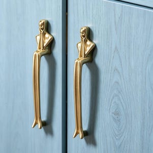 以下が含まれることがあります： 顔に手を当てて座っている様式化された人物の形をした、金色の金属製キャビネットハンドル2つ。人物の長い脚がハンドルのグリップ部分を形成しており、木目調のライトブルーのキャビネットの扉に垂直に取り付けられています。