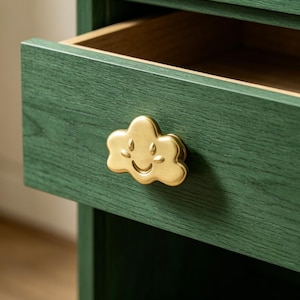 May include: A dark green wooden drawer featuring a gold-toned metal cabinet knob shaped like a smiling cloud with embossed facial features. The drawer is partially open, revealing a natural wood interior finish and a visible wood grain texture.