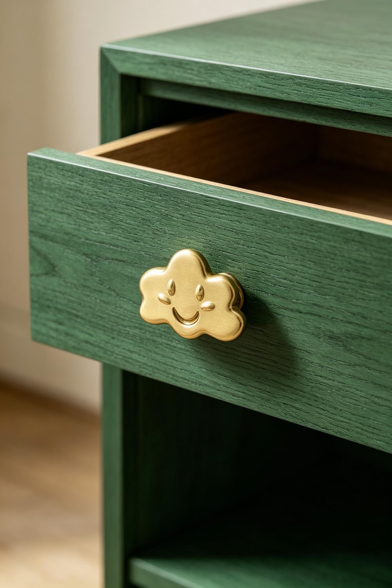 May include: A dark green wooden drawer featuring a gold-toned metal cabinet knob shaped like a smiling cloud with embossed facial features. The drawer is partially open, revealing a natural wood interior finish and a visible wood grain texture.
