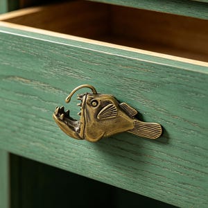 Puede incluir: Cajón de madera verde con un tirador en forma de pez de latón. El tirador es una escultura detallada de un pez con la boca abierta y una luz en la cabeza. El cajón está abierto, revelando un interior de madera.