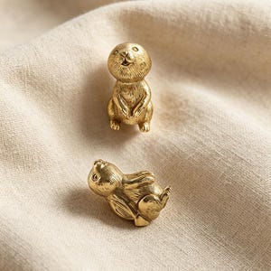 May include: Two small, metallic gold-toned figurines shaped like rabbits. One rabbit is depicted in a seated, upright position facing forward, while the second rabbit is shown lying down on its side. Both figures feature detailed, etched fur textures.