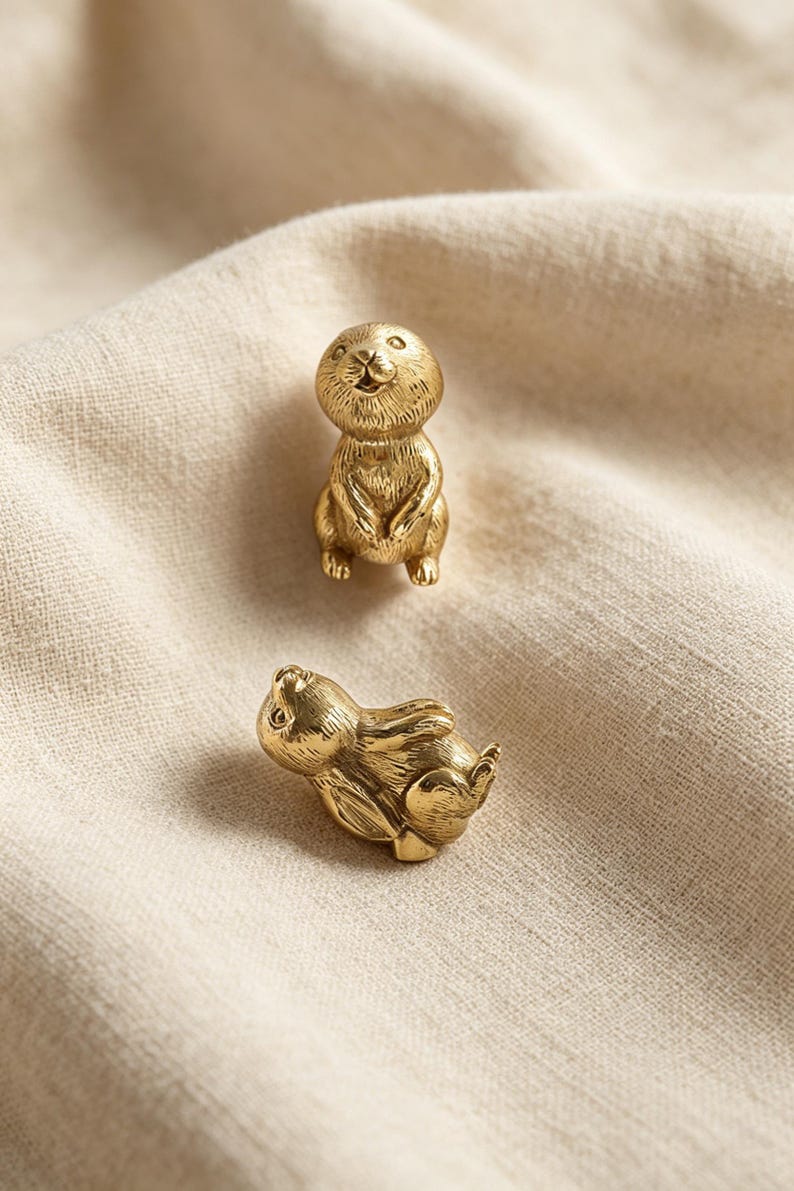 May include: Two small, metallic gold-toned figurines shaped like rabbits. One rabbit is depicted in a seated, upright position facing forward, while the second rabbit is shown lying down on its side. Both figures feature detailed, etched fur textures.