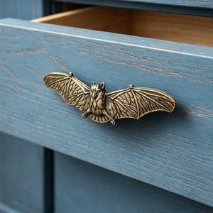 Könnte beinhalten: Ein messingfarbener Fledermaus-Griff ist an einer blauen Holzschublade befestigt. Die Flügel der Fledermaus sind ausgebreitet, und die Schublade ist teilweise geöffnet, wodurch ein Holzinnenraum sichtbar wird. Die Hardware verleiht den Möbeln eine einzigartige, gotische Note.