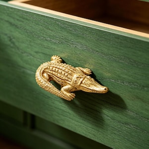 May include: A gold-toned metal drawer pull shaped like a detailed alligator, mounted on a dark green wooden drawer front with a visible wood grain texture. The drawer is slightly open, revealing the light-colored interior wood construction.