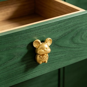 May include: A close-up of a green wooden drawer with a gold-coloured mouse-shaped knob. The drawer is open, revealing a light-coloured interior. The mouse knob has a small piece of cheese on its chest.