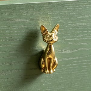 Op de afbeelding: Een gepolijste goudkleurige ladeknop in de vorm van een zittende kat met puntige oren en gedetailleerde gelaatstrekken. De katvormige handgreep is gemonteerd op een saliegroen houten ladefront met een zichtbare horizontale houtnerfstructuur.