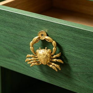 May include: A gold-colored crab-shaped drawer pull on a green wooden drawer. The drawer is open, revealing the interior. The crab handle has a coin detail. The wood grain is visible on the drawer front.
