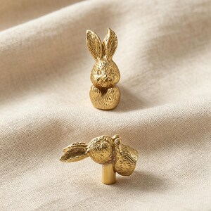 May include: Two gold-toned metal cabinet knobs shaped like small rabbits. One knob is shown from a front-facing perspective, while the other is positioned horizontally to reveal the cylindrical mounting post extending from the base of the rabbit figure.