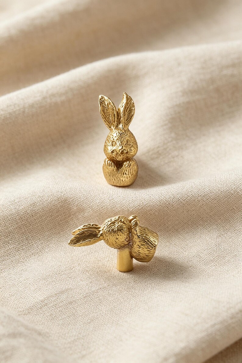 May include: Two gold-toned metal cabinet knobs shaped like small rabbits. One knob is shown from a front-facing perspective, while the other is positioned horizontally to reveal the cylindrical mounting post extending from the base of the rabbit figure.