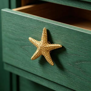 May include: A gold-colored starfish-shaped drawer pull is mounted on a dark green wooden drawer. The drawer is partially open, revealing a light brown interior. The starfish has a textured surface, adding a decorative touch to the furniture.