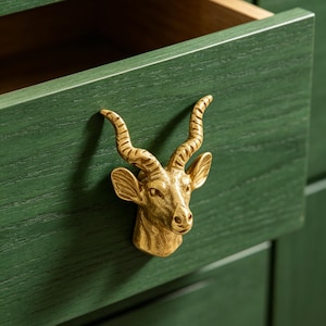 May include: A dark green wooden drawer featuring a metallic gold-toned cabinet pull shaped like the head of an antelope with curved, textured horns. The drawer is partially open, revealing a light-coloured interior wood finish and grain.