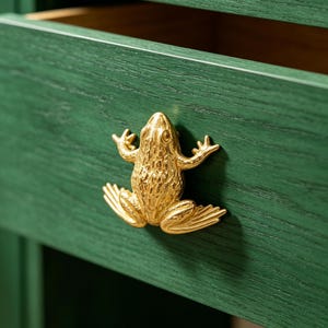May include: A dark green wooden drawer front featuring a textured gold-coloured metal frog-shaped knob. The drawer is slightly open, revealing a natural wood interior. The frog knob is centred on the front panel, showcasing detailed legs and skin.