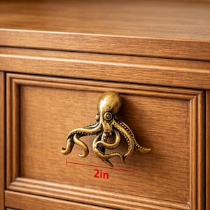 May include: A brass-toned metal cabinet knob shaped like an octopus with detailed tentacles and a rounded head. The knob is mounted on a wooden drawer front, and a red graphic line indicates a width measurement of 2 inches for the octopus.
